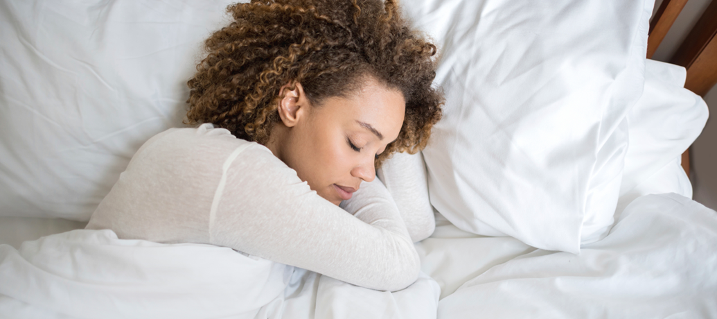 Woman with beautiful curly hair sleeps soundly on all-white bedding.
