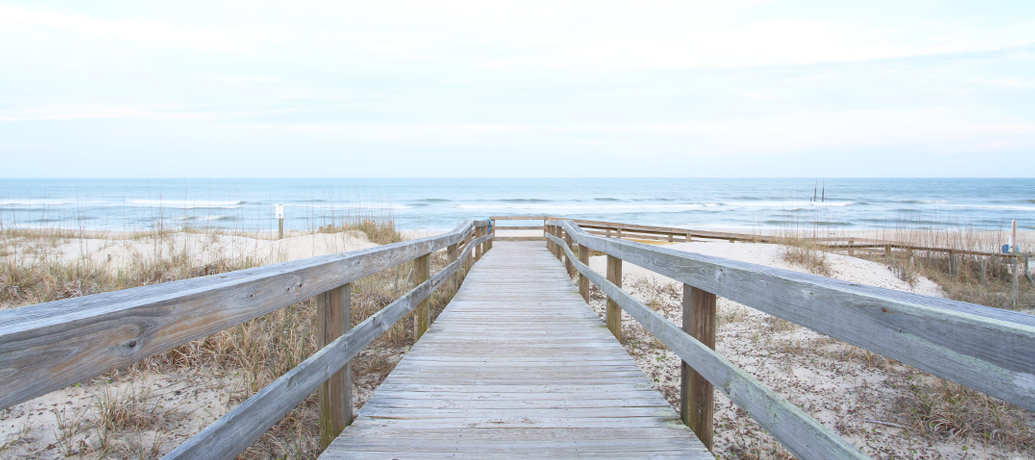 Beach boardwalk across sand dunes leads a view to the open blue water.