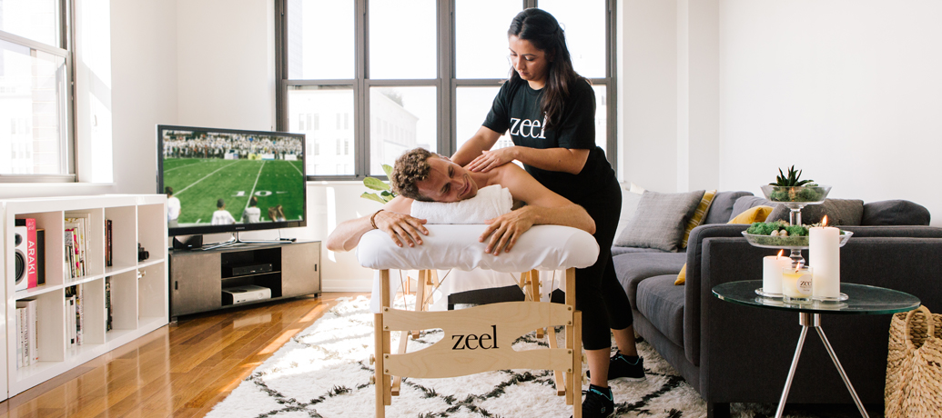 Man receives a massage in the comfort of his own apartment, while watching the big football game.