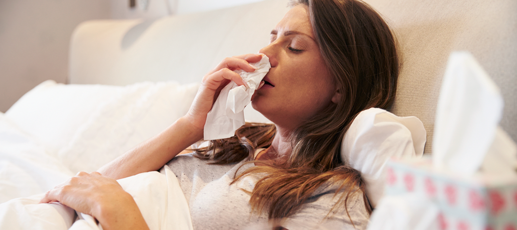 Woman lays under the covers in bed, taking a tissue to her nose in sickness.