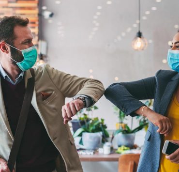 Two people bumping elbows while wearing PPE to safely greet each other during the pandemic.
