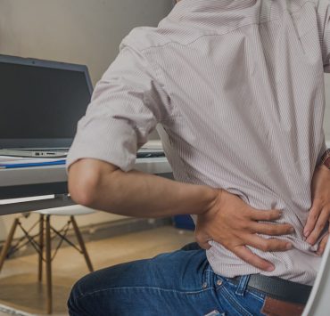 A man holing his lower back in pain as he sits at his desk since he forgot to to his upper body stretch for the day.