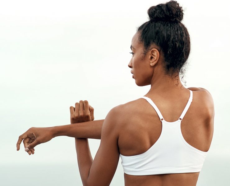 A woman srtanding outside stretching her arm and shoulder.