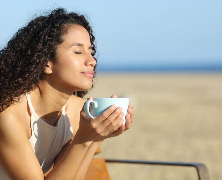 A woman having a drink on the beach as she prasctices mindfulness and takes the time to check in with herself.