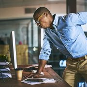 A man leaning over on his desk and holding his back in pain due to his poor spinal posture.