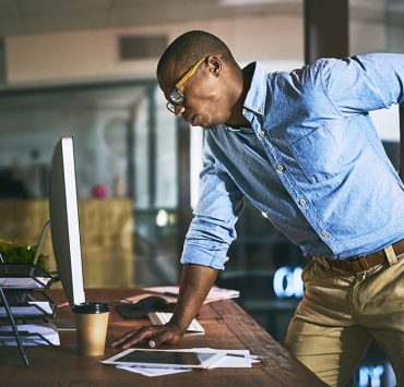 A man leaning over on his desk and holding his back in pain due to his poor spinal posture.