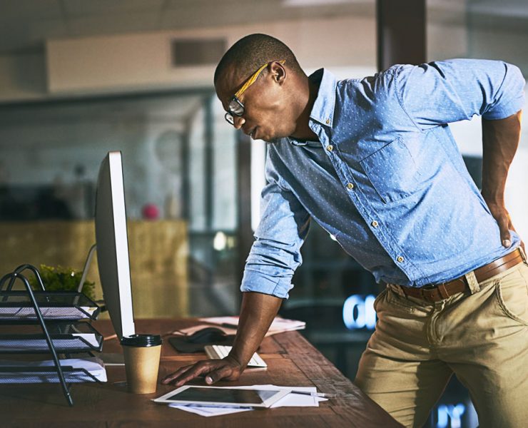 A man leaning over on his desk and holding his back in pain due to his poor spinal posture.